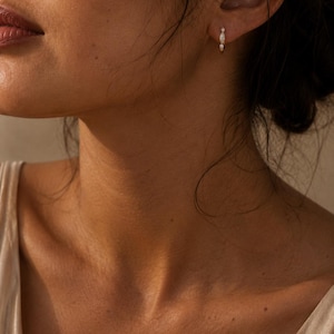 May include: A small gold hoop earring featuring a central marquise-cut white stone flanked by two tiny round clear stones. The earring is worn in the earlobe, showcasing a delicate metallic band and a trio of stones arranged in a vertical line.