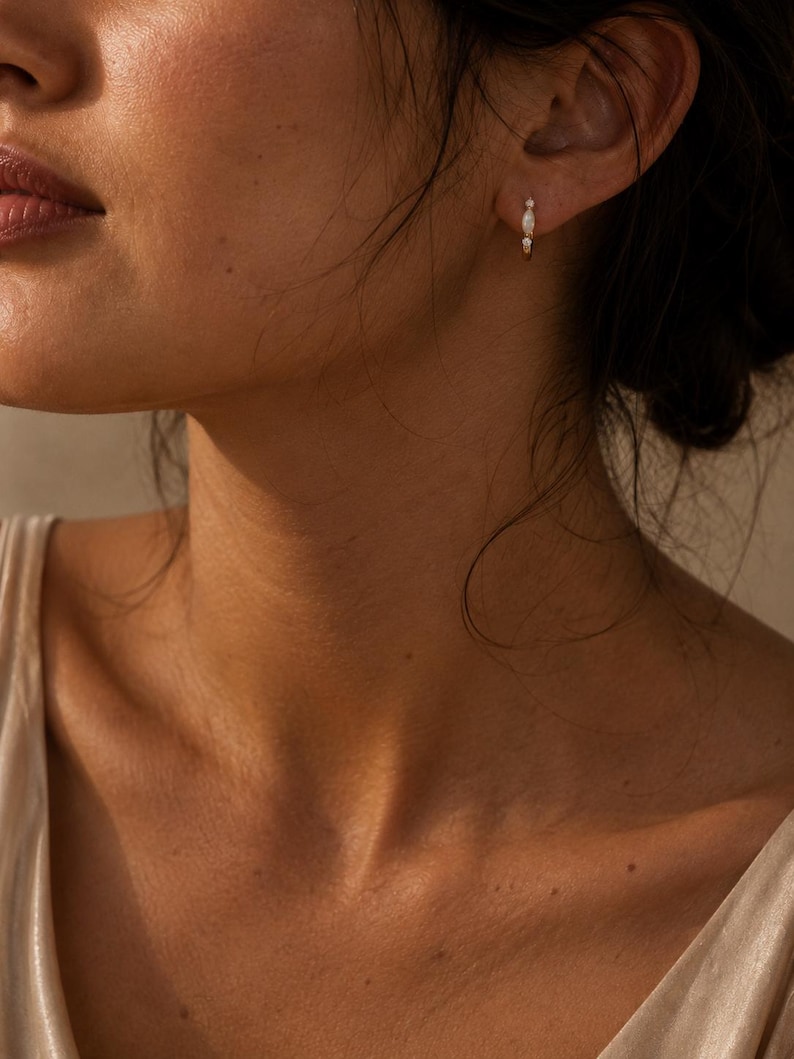 May include: A small gold hoop earring featuring a central marquise-cut white stone flanked by two tiny round clear stones. The earring is worn in the earlobe, showcasing a delicate metallic band and a trio of stones arranged in a vertical line.