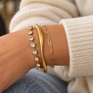 May include: A set of three gold-toned bracelets worn together. The first is a delicate chain featuring round, clear, bezel-set stones. The second is a smooth, solid, curved metal cuff. The third is a gold-toned chain with elongated oval links.