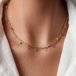May include: A gold-toned necklace featuring a twisted, textured chain. Small, dangling, multi-coloured faceted gemstones in shades of blue, pink, and clear are spaced evenly along the length of the chain, creating a delicate and shimmering appearance.