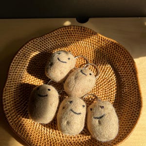 Könnte beinhalten: Sechs beige Filz-Kartoffel-Schlüsselanhänger mit lächelnden Gesichtern liegen auf einem gewebten braunen Korb. Jeder Schlüsselanhänger hat einen silbernen Ring. Der Korb ist rund und hat eine strukturierte Oberfläche.