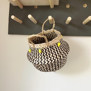 May include: A black and natural woven basket with yellow lemon accents hanging on a wall rack. The basket has a zig-zag pattern.