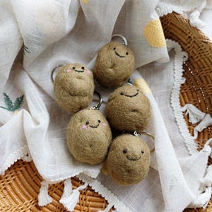May include: Five round, brown, needle-felted potato keychains with embroidered black eyes and smiling mouths. Two of the potato figures feature small, circular pink blush marks on their cheeks. Each keychain includes a silver metal ring attachment.
