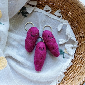 May include: Three magenta needle-felted wool keychains shaped like small, elongated tubers. Each keychain features a simple embroidered black thread face with two small dot eyes and a curved smile, attached to a circular silver metal keyring.