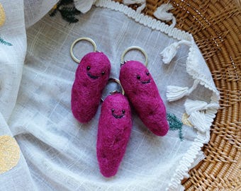 May include: Three magenta needle-felted wool keychains shaped like small, elongated tubers. Each keychain features a simple embroidered black thread face with two small dot eyes and a curved smile, attached to a circular silver metal keyring.
