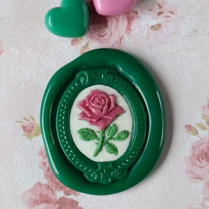 Puede incluir: Un sello de cera verde con un diseño de rosa rosa en un marco ovalado, acompañado de un adorno en forma de corazón verde y rosa. El fondo presenta un patrón floral.