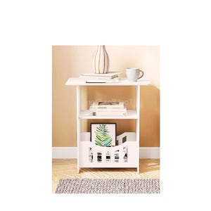 Może przedstawiać: Biały stolik nocny z trzema półkami. Na górnej półce stoi wazon w paski i książka. Na środkowej półce leżą dwie książki. Na dolnej półce znajduje się stojak na czasopisma z ramką na zdjęcia i czasopismami.