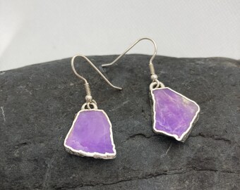 May include: A pair of silver earrings with purple gemstone accents. The earrings are made of a silver wire with a small loop at the top for hanging. The gemstones are irregular in shape and have a natural, raw look.