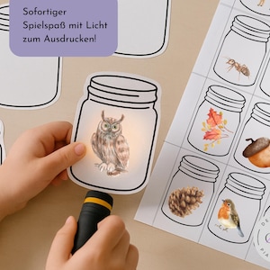 Può includere: Una mano di bambino tiene una torcia che illumina un'illustrazione di gufo all'interno di un ritaglio di barattolo. Sono visibili altri ritagli di barattoli e un foglio con varie illustrazioni a tema natura in barattoli. Il testo tedesco recita "Sofortiger Spielspaß mit Licht zum Ausdrucken!"