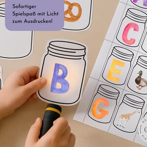 Peut inclure: Une main d'enfant illumine la lettre 'B' à l'intérieur d'une carte en forme de bocal avec une lampe de poche. L'image comprend d'autres cartes en forme de bocal avec des lettres et des illustrations. Le texte de l'image se lit : 'Amusement instantané avec la lumière à imprimer !'