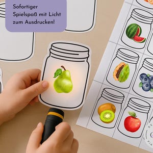May include: A child's hand holds a pear-shaped card illuminated by a flashlight, part of a set of fruit-themed cards in mason jars. Other cards feature watermelon, apple, papaya, blueberries, kiwi, apple, and pineapple. Text in German reads "Immediate fun with light to print!"