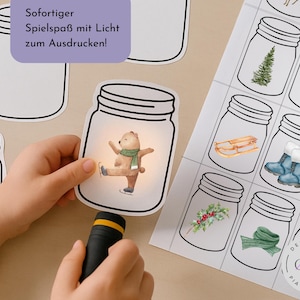 Könnte beinhalten: Ein Kinderhand hält eine Taschenlampe, die eine Bärenillustration in einem Einmachglas beleuchtet. Der Bär trägt einen grünen Schal und fährt Schlittschuh. Weitere Gläser enthalten winterliche Bilder wie einen Schneemann und einen Weihnachtsbaum.