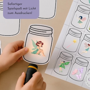 Op de afbeelding: Een kinderhand houdt een zaklamp vast die een fee in een glazen pot-uitsparing verlicht. Andere potuitsparingen met feeën en magische voorwerpen liggen op een wit oppervlak. Tekst in het Duits.