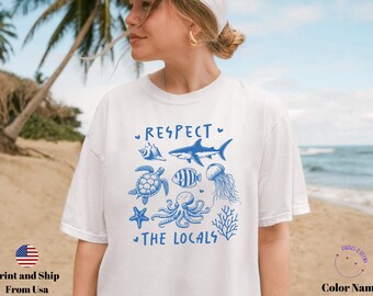May include: White t-shirt with a blue graphic of various sea creatures, including a shark, turtle, and octopus. The text "RESPECT THE LOCALS" is printed above and below the illustrations. The shirt is worn by a person on a beach.