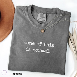 May include: A grey Comfort Colors t-shirt with the slogan "none of this is normal." A gold pendant necklace is on the shirt. The shirt is labelled "Pepper" at the bottom.