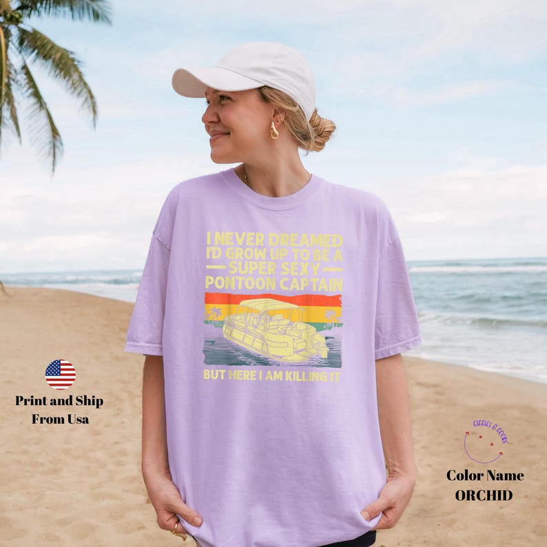 K&ouml;nnte beinhalten: Orchideenfarbenes T-Shirt mit Retro-Design, das ein Pontonboot und den Text "I never dreamed I'd grow up to be a super sexy pontoon captain, but here I am killing it" zeigt. Das Shirt wird von einer Person am Strand getragen.