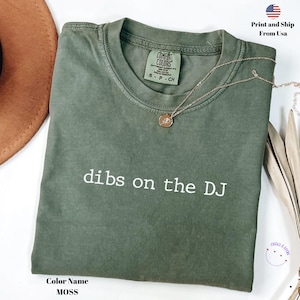 Könnte beinhalten: Ein moosgrünes T-Shirt mit dem weißen Aufdruck "dibs on the DJ". Das Shirt hat einen Rundhalsausschnitt und kurze Ärmel. Eine goldene Halskette mit Anhänger liegt auf dem Shirt. Das Shirt ist mit "Comfort Colors" gekennzeichnet.