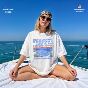 K&ouml;nnte beinhalten: Wei&szlig;es T-Shirt mit dem Text "I NEVER DREAMED I'D GROW UP TO BE A -SUPER SEXY- PONTOON CAPTAIN BUT HERE I AM KILLING IT" in Blau und einer Grafik eines Pontonboots. Das T-Shirt wird von einer Person auf einem Boot getragen.