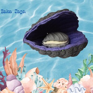 Puede incluir: Una concha de almeja de crochet de color morado y gris con una almeja gris y blanca en su interior. La concha de almeja está sobre un fondo azul con algas marinas y coral.