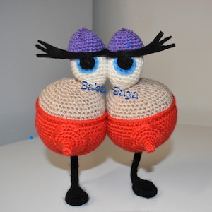 Peut inclure: Une figurine fantaisiste faite à la main au crochet, avec deux grands yeux ronds, chacun avec un iris bleu et une pupille noire. Le corps est beige avec une base rouge, des jambes noires et un chapeau violet. Le texte "Baba Jaga" est visible.