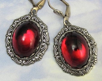 May include: A pair of ornate earrings featuring large, oval, ruby-red glass stones set in silver-toned filigree frames. The earrings have lever-back closures and a vintage aesthetic, suitable for formal occasions or adding a touch of elegance.