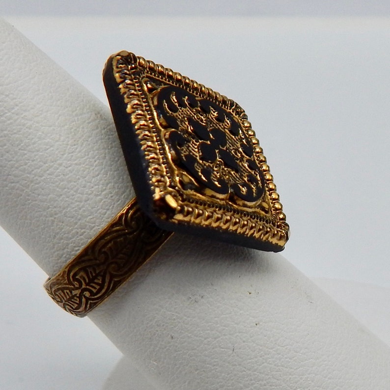 May include: Gold ring with a black square-shaped stone featuring an intricate gold design.