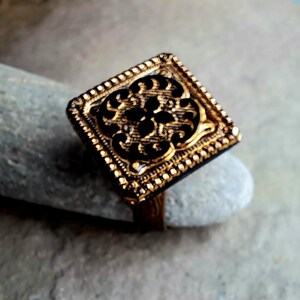 May include: A gold ring with a square-shaped design featuring a detailed, intricate pattern. The ring has a textured border.