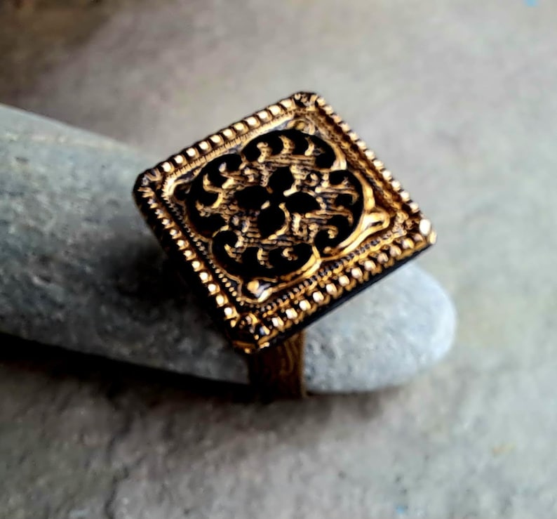 May include: A gold ring with a square-shaped design featuring a detailed, intricate pattern. The ring has a textured border.