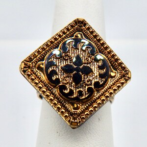 May include: A gold ring with a black enamel square design featuring a floral pattern. The ring has a textured border.