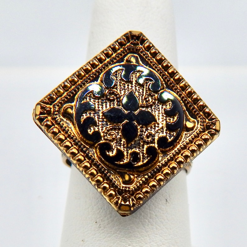 May include: A gold ring with a black enamel square design featuring a floral pattern. The ring has a textured border.