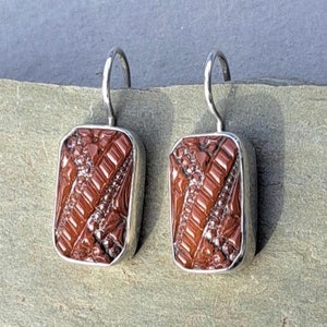 Puede incluir: Un par de pendientes de plata con piedras rectangulares de vidrio marrón. Las piedras tienen un patrón texturizado y geométrico.