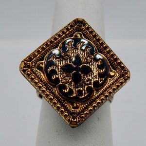 May include: A gold ring with a square black and gold design. The design features a black enamel floral pattern.