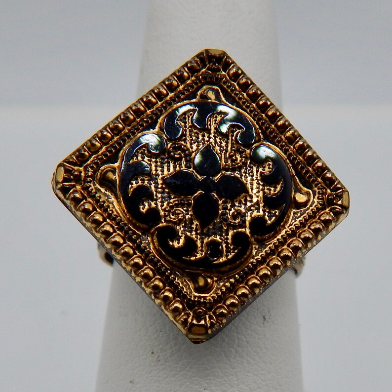 May include: A gold ring with a square black and gold design. The design features a black enamel floral pattern.