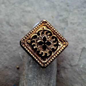 May include: A gold-colored square button with a black, intricate floral design. The button has a raised, textured border.