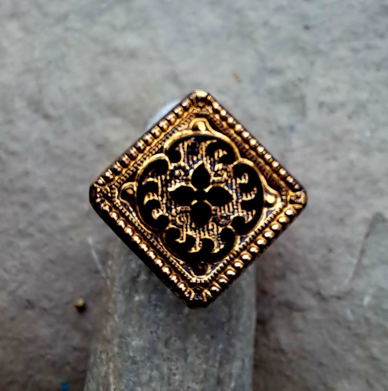 May include: A gold-colored square button with a black, intricate floral design. The button has a raised, textured border.