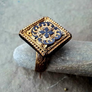 May include: A gold ring with a square black stone set in a gold frame. The stone has a detailed floral design.