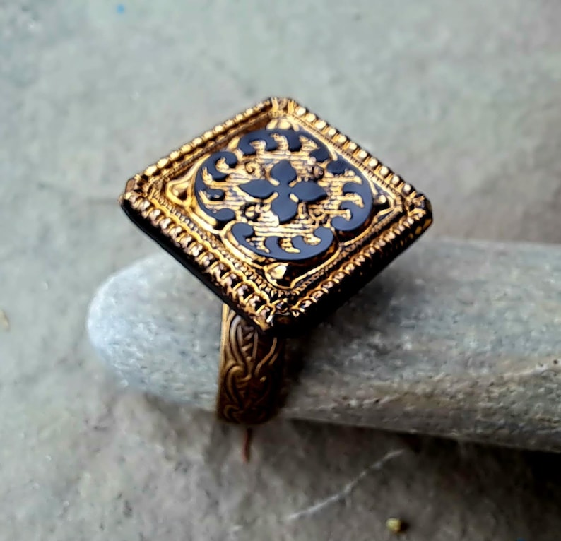 May include: A gold ring with a square black stone set in a gold frame. The stone has a detailed floral design.