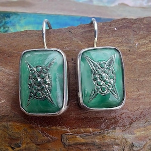 Puede incluir: Un par de pendientes rectangulares con marco plateado. Cada pendiente presenta un fondo verde claro con un diseño de estrella plateada en el centro. Los pendientes tienen cierre de gancho.