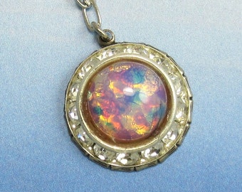 May include: A gold-tone pendant necklace with a round, iridescent opal-like stone set in a bezel setting. The stone is surrounded by clear rhinestones.