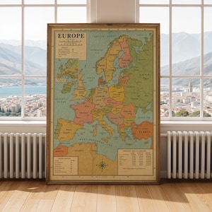 Op de afbeelding: Een vintage wandkaart van Europa in een donkerhouten lijst, uitgevoerd in gedempte tinten groen, geel en oranje. De kaart toont landsgrenzen, grote steden, een legenda, een schaal in mijlen en een tabel met geschatte oppervlaktes en bevolking.