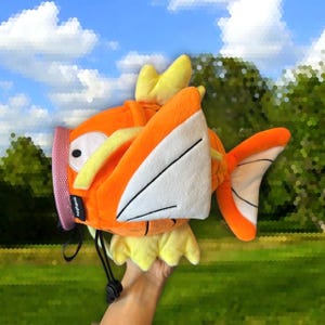 Könnte beinhalten: Orangefarbenes und gelbes Plüschtier in Form eines Fisches mit einer weißen Flosse. Das Spielzeug hat einen Kordelverschluss und ist ein Charakter aus dem Pokemon-Franchise.