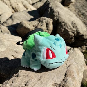 Puede incluir: Un juguete de peluche verde y azul con forma de personaje Pokémon, Bulbasaur. El juguete tiene ojos rojos y una hoja verde en la espalda. El juguete está sentado en una roca.