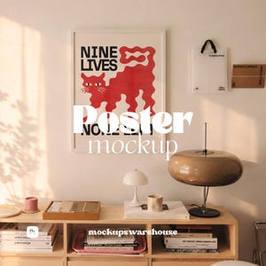 May include: A framed poster featuring a red cat illustration and the text NINE LIVES in black, displayed on a wall above a wooden shelf containing books, a small white lamp, a brown glass lamp, and a tray with various items.