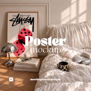 May include: A vertical rectangular poster in a light wood frame featuring a black Stussy logo above two red dice with black dots. The poster is displayed next to a chrome mushroom lamp, a stack of three books, and a white textured bedspread.