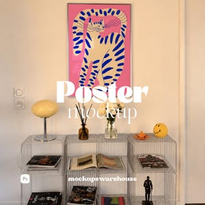 May include: A rectangular wall poster featuring a stylized cream-colored cat with blue stripes against a vibrant pink background, displayed in a silver frame above a wire grid shelving unit containing books, a lamp, a vase of flowers, and decor.