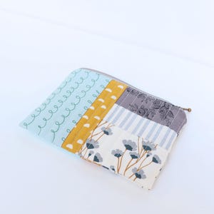 May include: A patchwork zipped pouch with various fabric patterns. It features sections of light blue with white swirls, mustard yellow, grey floral, and cream with blue flowers. The zip is silver in colour.