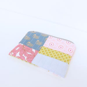 May include: A patchwork zipped pouch with a variety of fabric patterns. The design includes sections of pink leaves, blue with gold accents, pink circles, yellow floral, and white polka dots. The pouch has a grey zip.