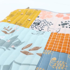 May include: Close-up of a patchwork fabric with various patterns and colours. The design includes squares of yellow, peach, light blue, and orange, each with different printed motifs like foxes, flowers, and geometric shapes. The fabric is quilted with vertical stitching.