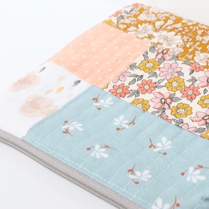 May include: A patchwork fabric item with a grey zip. The fabric features floral and patterned sections in peach, light blue, and mustard yellow. Visible stitching adds to the handcrafted feel. The item is likely a pouch or small bag.