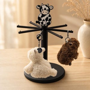 May include: A black metal rotating display stand featuring a cow-shaped topper. The stand has multiple horizontal arms for hanging items. Two plush keychains, one white and one brown, are suspended from the arms of the circular base stand.